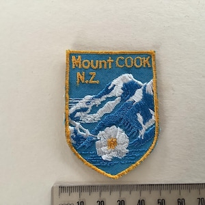 Puede incluir: Parche bordado con forma de escudo que muestra "Mount COOK N.Z." en letras doradas. El diseño representa una cordillera azul y blanca y una flor blanca. El parche tiene un borde dorado.