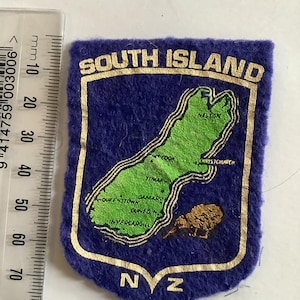 May include: A blue felt patch with a gold border and the words "SOUTH ISLAND" at the top. The patch features a green map of the South Island of New Zealand, with the letters "NZ" at the bottom.