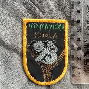 May include: A black patch with a gold border featuring a koala bear design. The patch has the word "KOALA" in gold lettering, with green text above. The koala bears are gray and white, sitting on a brown tree branch.
