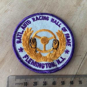 Peut inclure: Écusson circulaire avec une bordure violette et un centre bleu clair. L'écusson présente un volant doré avec des mains, entouré d'une couronne de laurier dorée. Le texte "NATL AUTO RACING HALL OF FAME FLEMINGTON, N.J." est brodé sur l'écusson.