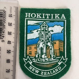 May include: A green felt patch featuring the text "HOKITIKA" at the top and "NEW ZEALAND" on a banner at the bottom. The patch has a rectangular design with rounded corners. The center depicts a detailed illustration of a building with a clock tower, set against a blue sky.
