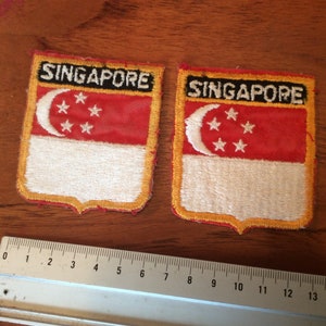 Puede incluir: Dos parches bordados en rojo, blanco y dorado con el texto "SINGAPORE" y una luna creciente con cinco estrellas.