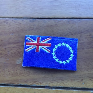 Pode incluir: Um remendo bordado azul com a bandeira das Ilhas Cook. A bandeira apresenta uma Union Jack no canto superior esquerdo e um círculo de estrelas brancas no canto inferior direito.