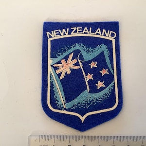 May include: A blue felt patch with a white border and the words "NEW ZEALAND" at the top. The patch features two flags with the New Zealand flag design. The flags are light blue with a white border and a pink and white design.