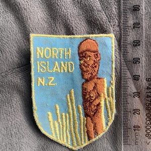 May include: A shield-shaped patch with the text "NORTH ISLAND N.Z." in yellow on a light blue background. The patch features an embroidered brown figure and yellow grass-like elements. A ruler is next to the patch.