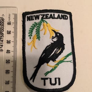 May include: Embroidered patch featuring a black and white bird perched on a green branch with yellow flowers. The patch has the words "NEW ZEALAND" and "TUI" in black text on a white background, with a black border.