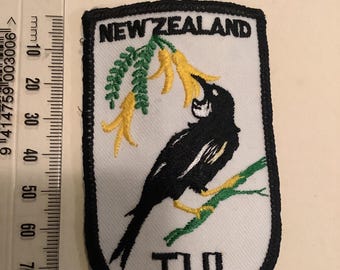 Op de afbeelding: Geborduurde patch met een zwart-witte vogel op een groene tak met gele bloemen. De patch heeft de woorden "NEW ZEALAND" en "TUI" in zwarte tekst op een witte achtergrond, met een zwarte rand.
