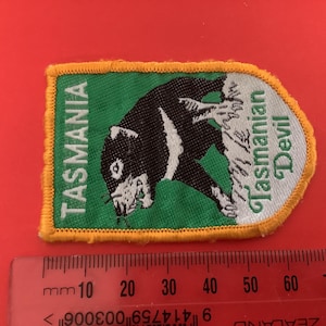 Pode incluir: Um patch retangular com fundo verde e borda amarela. O patch apresenta uma ilustração em preto e branco do demônio da Tasmânia e o texto "TASMANIA" e "Tasmanian Devil".
