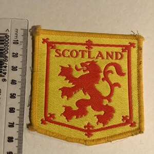 May include: A yellow and red embroidered patch featuring the word "SCOTLAND" and a red lion rampant emblem. The patch has a yellow border and is approximately 3 inches wide.