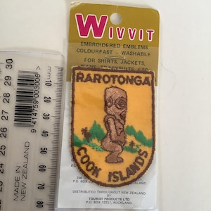 May include: Embroidered patch from Rarotonga, Cook Islands, featuring a brown tiki figure and palm trees on a yellow background. The patch is labeled "Wivvit" and is designed for shirts, jackets, and jeans.