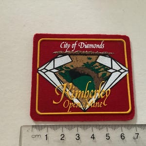 May include: Red fabric patch with a yellow border. The patch features a white diamond with a green and brown image inside. The text "City of Diamonds Kimberley Open Mine" is printed in yellow.