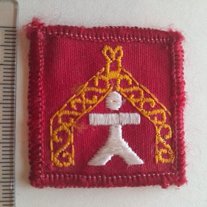May include: A square, red fabric patch with a white and gold embroidered design. The design features a stylized figure under an arch. The patch is approximately 1.5 inches square, with a red stitched border.