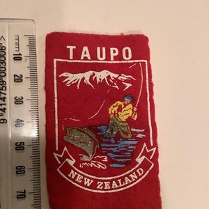 May include: A rectangular red felt patch with white text and a graphic of a fisherman. The text at the top reads "TAUPO", and the text at the bottom reads "NEW ZEALAND". The graphic depicts a fisherman catching a fish.