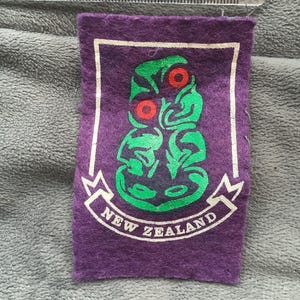 May include: A purple felt patch with a white border and the words "NEW ZEALAND". The patch features a green and red graphic of a Maori design. The patch measures approximately 2 inches wide.