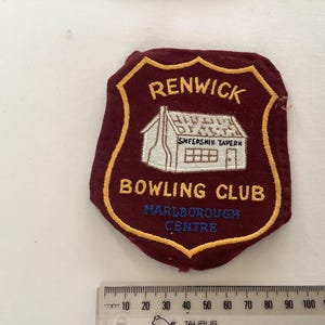 Può includere: Toppa ricamata bordeaux e oro del Renwick Bowling Club. La toppa presenta il testo "RENWICK BOWLING CLUB MARLBOROUGH CENTRE" e un'illustrazione di un edificio con l'etichetta "SHEEPSKIN TAVERN".