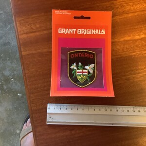 Op de afbeelding: Een geborduurde patch met de tekst "ONTARIO" in rode letters, in de kleuren rood, wit en blauw. De patch is op een paarse achtergrond en is omgeven door een witte rand. De patch zit in een plastic verpakking met de tekst "GRANT ORIGINALS" erop gedrukt.