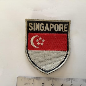 Puede incluir: Un parche bordado en rojo, blanco y plateado con la palabra "SINGAPORE" en letras negras y el diseño de la bandera de Singapur.