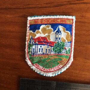May include: A white, blue, red, and gold embroidered patch with the text "WIESKIRCHE WALLFAHRT SORT" and a depiction of a church building.
