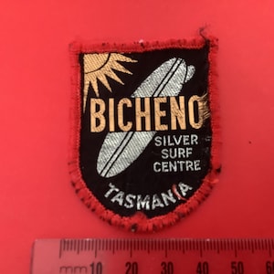 Peut inclure: Écusson noir, rouge et doré avec le texte "Bicheno Silver Surf Centre Tasmania". L'écusson présente un soleil et une planche de surf argentée. L'écusson est sur un fond rouge.