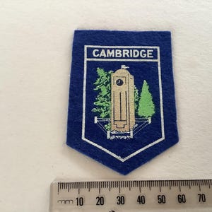 May include: A blue felt patch with a white border and a shield shape. The patch features the word "CAMBRIDGE" in white at the top. Below is a beige clock tower with green trees. A ruler is at the bottom.