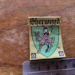 Może przedstawiać: Złota naszywka z tkaniny z napisem "Sherwood" czarnymi literami. Naszywka przedstawia zieloną tarczę z kreskówkowym obrazkiem osoby jeżdżącej konno. Tarcza ma dwa małe fioletowe i złote kółka po obu stronach.