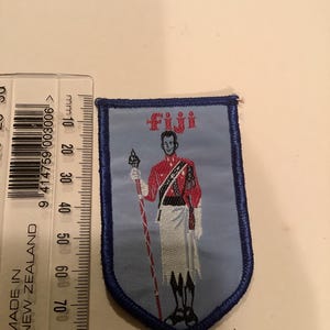 May include: A light blue patch with a shield shape and a dark blue border. The patch features a figure in a red jacket and white skirt holding a spear. The word "Fiji" is at the top in red letters. A ruler is on the left side of the patch.