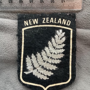 May include: A black felt patch with a shield shape, featuring a silver fern design and the words "NEW ZEALAND" in gold. The patch has a gold border and is approximately 2 inches wide.