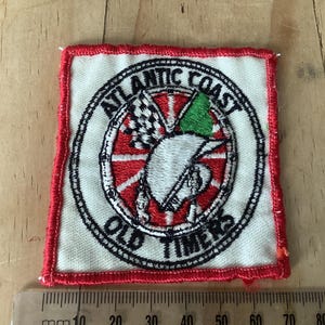 May include: Embroidered patch with a red border and a circular design. The design features the text "ATLANTIC COAST OLD TIMERS" around a central image of a white bird, a checkered flag, and a green triangle. The patch is square.