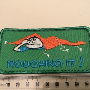 Może przedstawiać: Haftowana naszywka z zielonym tłem i zieloną obwódką. Naszywka przedstawia postać z kreskówki z pomarańczowymi włosami i niebieską koszulą, zasłaniającą oczy. Tekst "ROUGHING IT!" jest wyhaftowany na niebiesko.