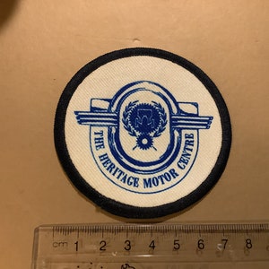Puede incluir: Un parche bordado blanco y azul con borde negro. El parche presenta un logotipo con el texto "The Heritage Motor Centre".
