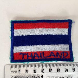May include: Embroidered patch featuring the flag of Thailand. The rectangular patch has horizontal stripes of red, white, and blue. The word "THAILAND" is embroidered in red at the bottom. The patch is approximately 5 cm wide.