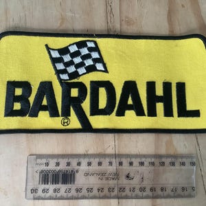 Peut inclure: Écusson brodé jaune avec le mot "BARDAHL" en noir et un drapeau à damier. Une règle en dessous indique les mesures en centimètres. Le patch mesure environ 15 cm de long.