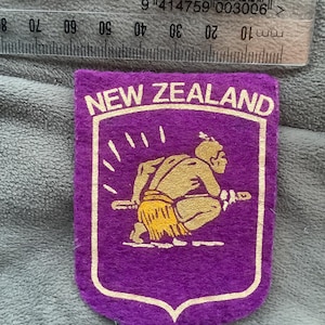 May include: A purple felt patch with the words "NEW ZEALAND" in white lettering. The patch features a gold-colored illustration of a person in a squatting position, holding a staff. The patch is shield-shaped.