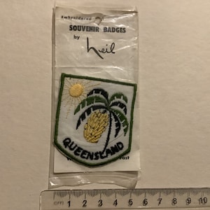 Op de afbeelding: Gestikt souvenirbadge met een groene palmboom met een gele ananas en een gele zon op een witte achtergrond. Het badge is omgeven door een groene rand met de tekst "QUEENSLAND" in zwarte letters. Het badge is verpakt in een doorzichtige plastic zak met een wit label waarop "Embroidered Souvenir Badges by Heil" staat.