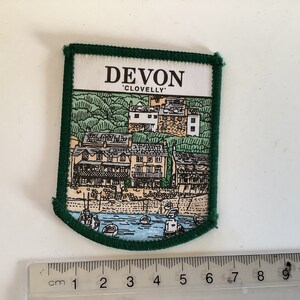May include: A green embroidered patch with a white background featuring a drawing of a village with the text "DEVON 'CLOVELLY'".