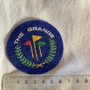 May include: A blue felt patch with the text "THE GRANGE" in white lettering around a green wreath with three gold golf clubs and red, yellow, and green flags.