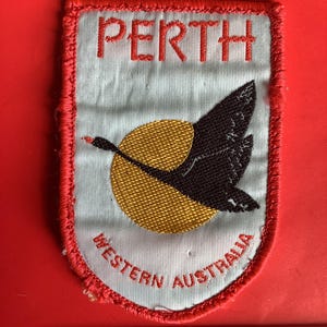 Op de afbeelding: Geborduurde patch met een zwarte zwaan in vlucht tegen een gele zon, met het woord "PERTH" erboven en "WESTERN AUSTRALIA" eronder. De patch heeft een rode rand en een lichtblauwe achtergrond.