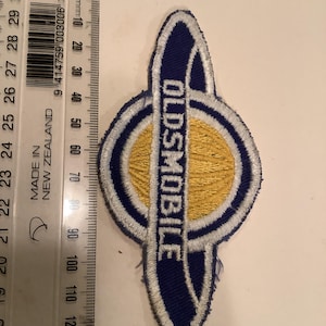 May include: Embroidered patch with the word "OLDSMOBILE" in white on a navy blue background. The patch has a gold center and a white border. The patch measures approximately 7.6 cm in length.