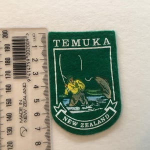 May include: Green felt patch with the word "TEMUKA" at the top and "NEW ZEALAND" at the bottom. The patch features an illustration of a fisherman and a fish. The patch is approximately 7.6 cm long.