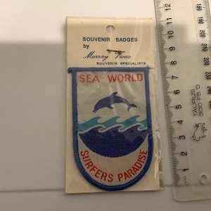 Peut inclure: Un écusson brodé bleu et blanc avec un dauphin sautant au-dessus des vagues. L'écusson porte l'inscription "Sea World" et "Surfers Paradise" en rouge et blanc. L'écusson est sur une carte blanche avec le texte "Souvenir Badges by Murray Views Souvenir Specialists".