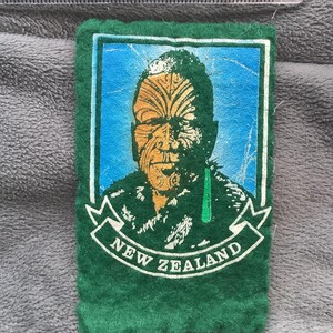 May include: A green felt patch featuring a stylized portrait of a Maori person with traditional facial tattoos, set against a blue background. The patch has the words "NEW ZEALAND" in a white banner.