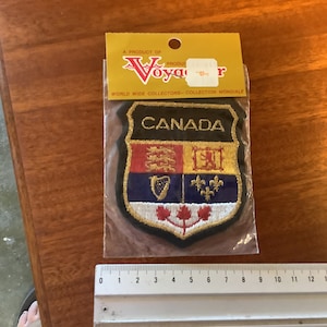 Könnte beinhalten: Ein gestickter schwarzer und goldener Aufnäher mit dem kanadischen Wappen. Der Aufnäher ist mit "CANADA" beschriftet und in einer durchsichtigen Plastiktüte verpackt, die ein gelbes und rotes Etikett mit der Aufschrift "Ein Produkt von Voyager Weltweite Sammler - Collection Mondiale" trägt.