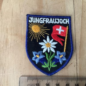Puede incluir: Parche bordado con borde azul y fondo negro. El parche presenta el texto "JUNGFRAUJOCH", un sol amarillo, una bandera roja con una cruz blanca, una flor blanca y flores azules. Una regla está en la parte inferior.