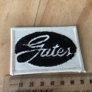 Peut inclure: Un écusson rectangulaire blanc avec un motif ovale noir et le mot "Gates" brodé en écriture blanche. L'écusson est posé sur une surface en bois à côté d'une règle.