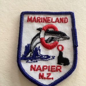 Puede incluir: Parche bordado con fondo blanco, borde azul y el texto "Marineland Napier N.Z.". Una orca en blanco y negro salta a través de un salvavidas rojo. También se representa una foca negra.