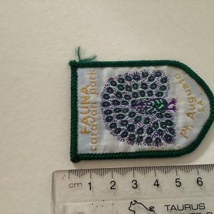 Puede incluir: Un parche bordado verde y blanco con el texto "FAUNA Garden Park" y "Ph. Augusta's" en letras doradas. Un diseño de pavo real está en el centro del parche.