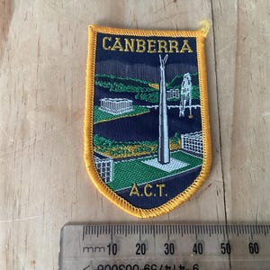 Puede incluir: Un parche en forma de escudo con borde dorado y la palabra "CANBERRA" en letras doradas. El parche presenta un paisaje con una estructura alta y el texto "A.C.T."