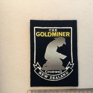 May include: A navy blue patch featuring a silver silhouette of a gold miner, with the text "THE GOLDMINER" in yellow and "Cromwell New Zealand" in white. The patch has a white border and is rectangular in shape.