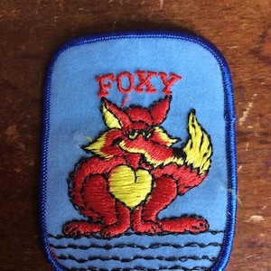May include: A blue felt patch with a red fox holding a yellow heart. The fox is embroidered and the patch has a blue border. The text "FOXY" is embroidered in red above the fox.