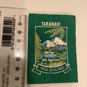 May include: A green souvenir with an embroidered image of Mount Egmont in New Zealand. The text "TARANAKI" and "NEW ZEALAND" are displayed. A ruler is placed next to the souvenir, showing measurements in millimeters.
